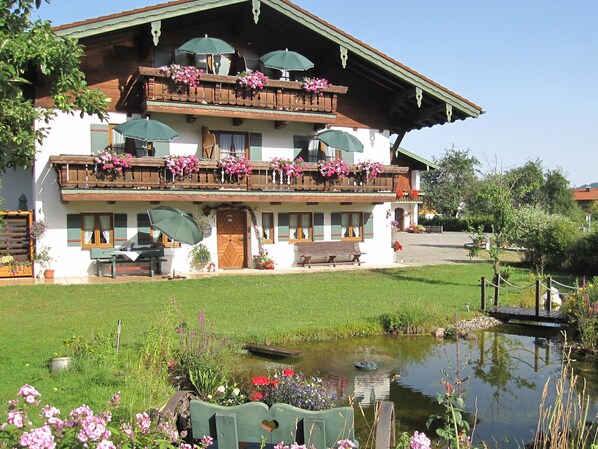 A two-story building with balconies adorned with flowers, a pond, and a bench in front.