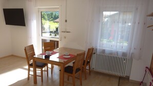 Un coin repas avec une table et des chaises en bois, un téléviseur fixé au mur et une vue sur l’extérieur par une fenêtre.