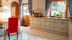 A kitchen with a wooden dining table, a red chair, white cabinets, a microwave, and a window with curtains. - Vacation apartment no. 1 Watzmann, 70sqm for 2 persons (Berchtesgaden)