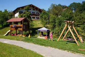 Explore the tranquil play area adjacent to our chalet, featuring a wooden playhouse, swing set, and tent-like structure amidst lush greenery.