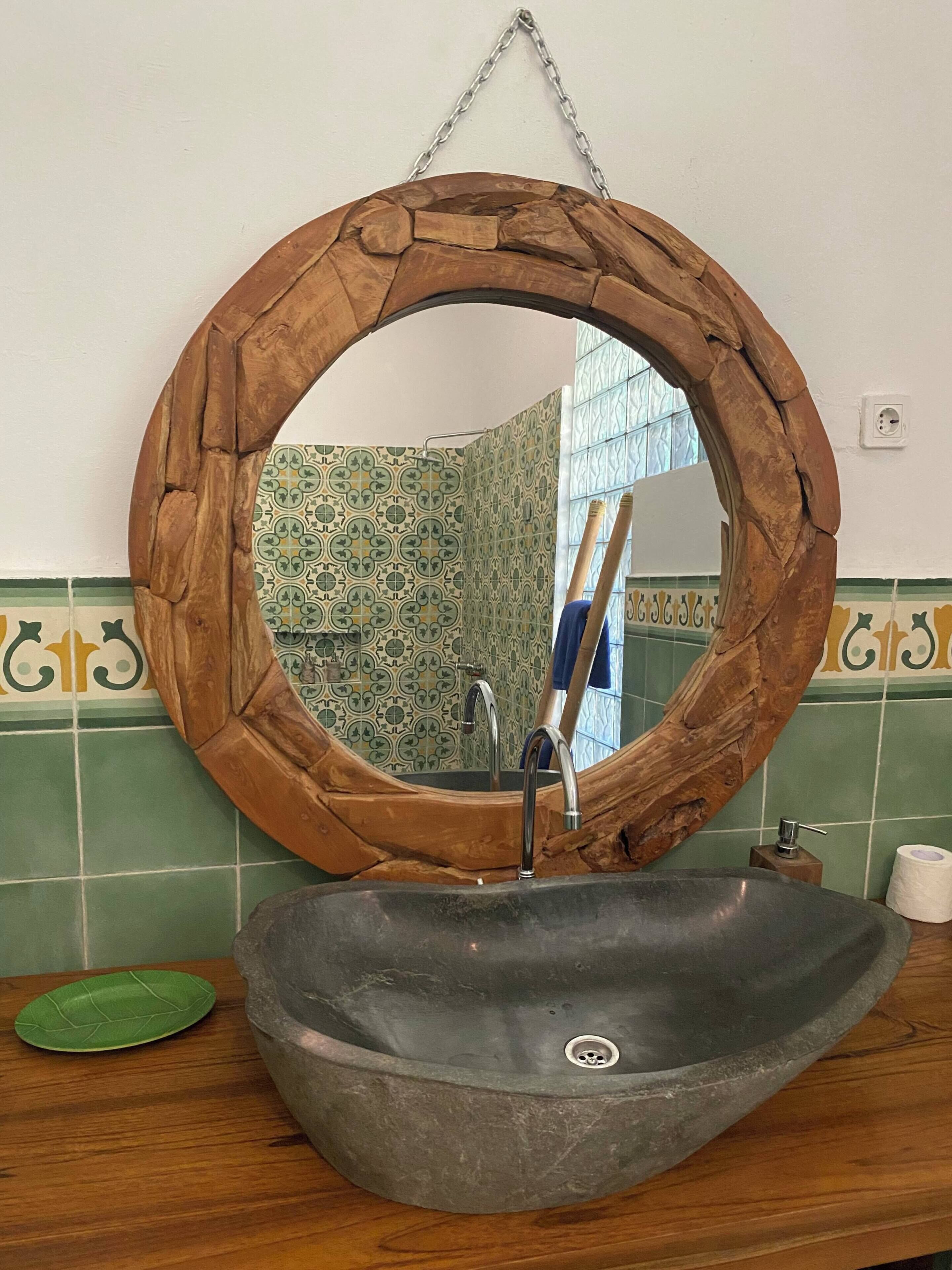 A bathroom with a round wooden-framed mirror, a stone sink, and green patterned tiles.