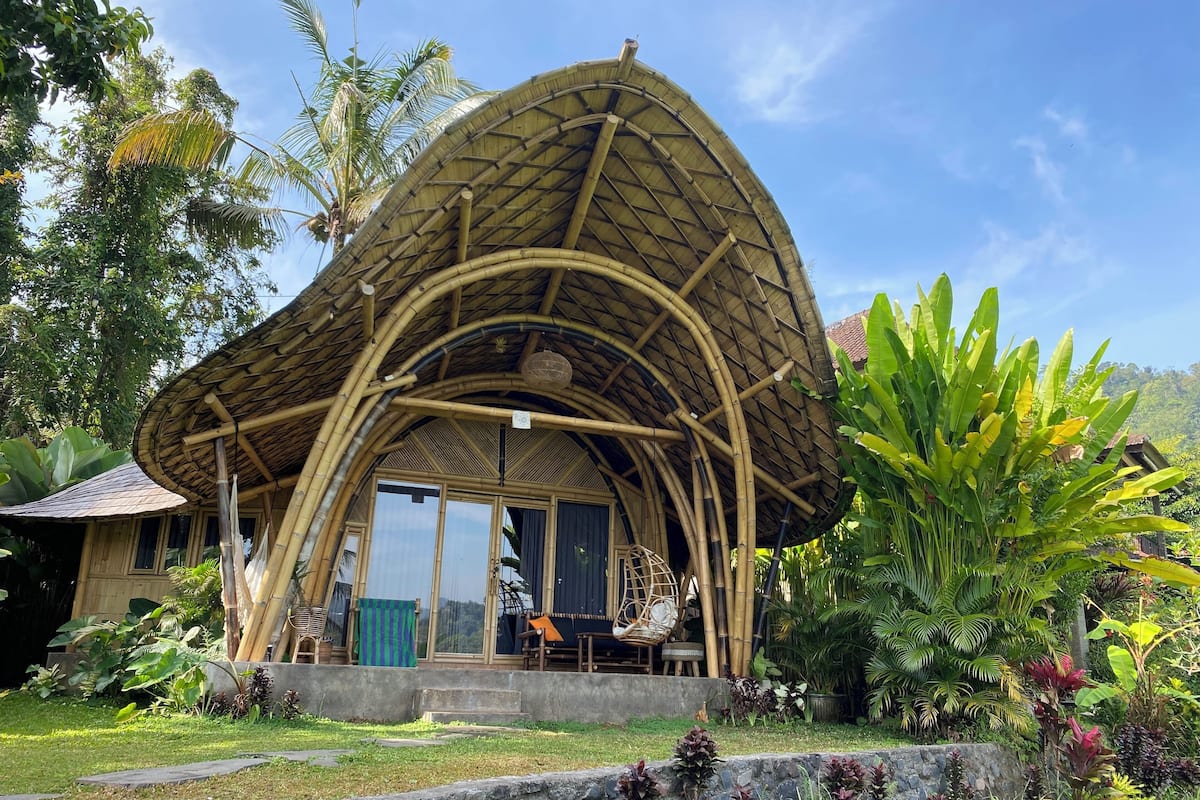 A bamboo structure with a curved roof, surrounded by lush greenery and a stone wall.
