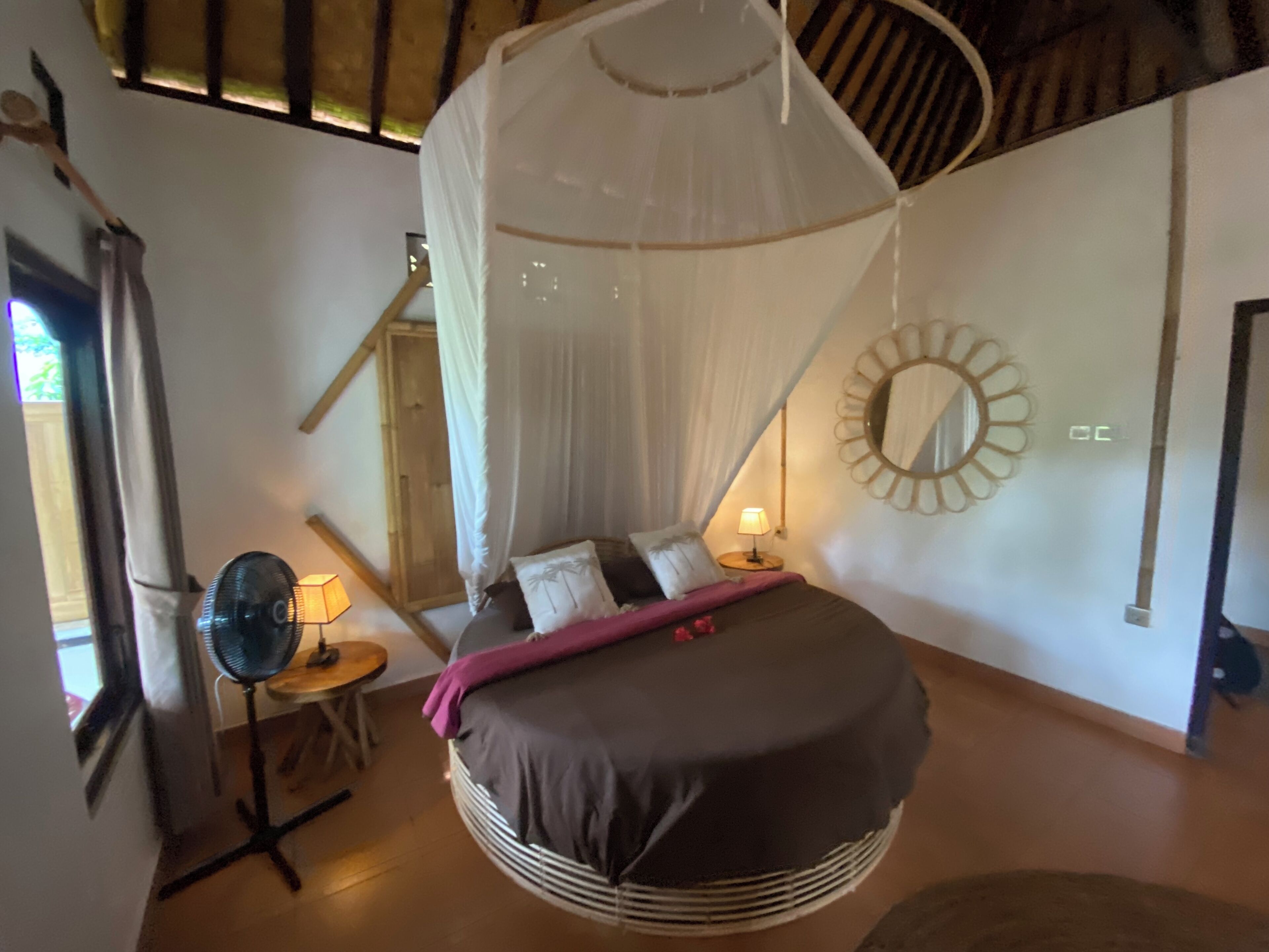 A bedroom with a bed under a canopy, a round mirror, a bedside table with a lamp, and a fan.