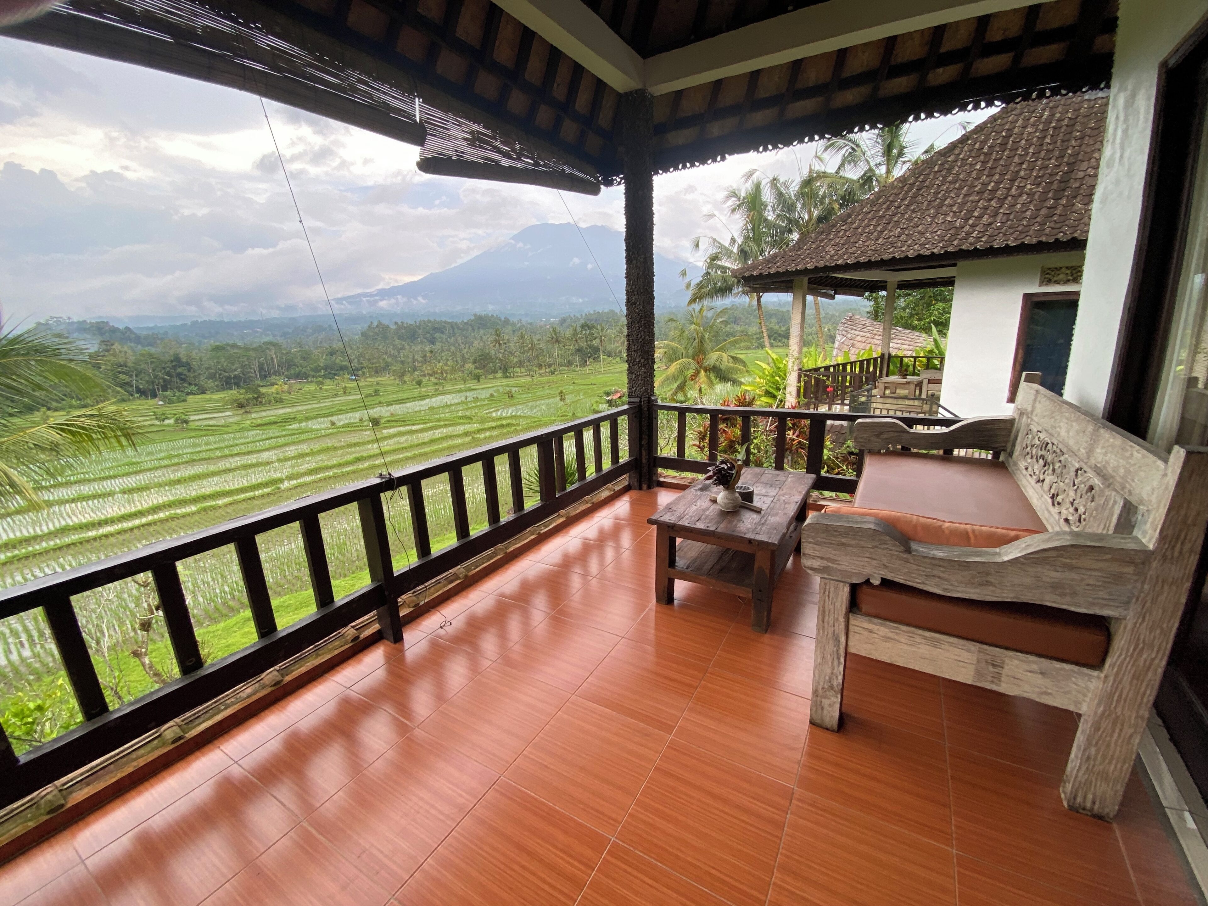 A balcony with a wooden bench and table, overlooking a lush green landscape with terraced fields and a mountain in the distance.