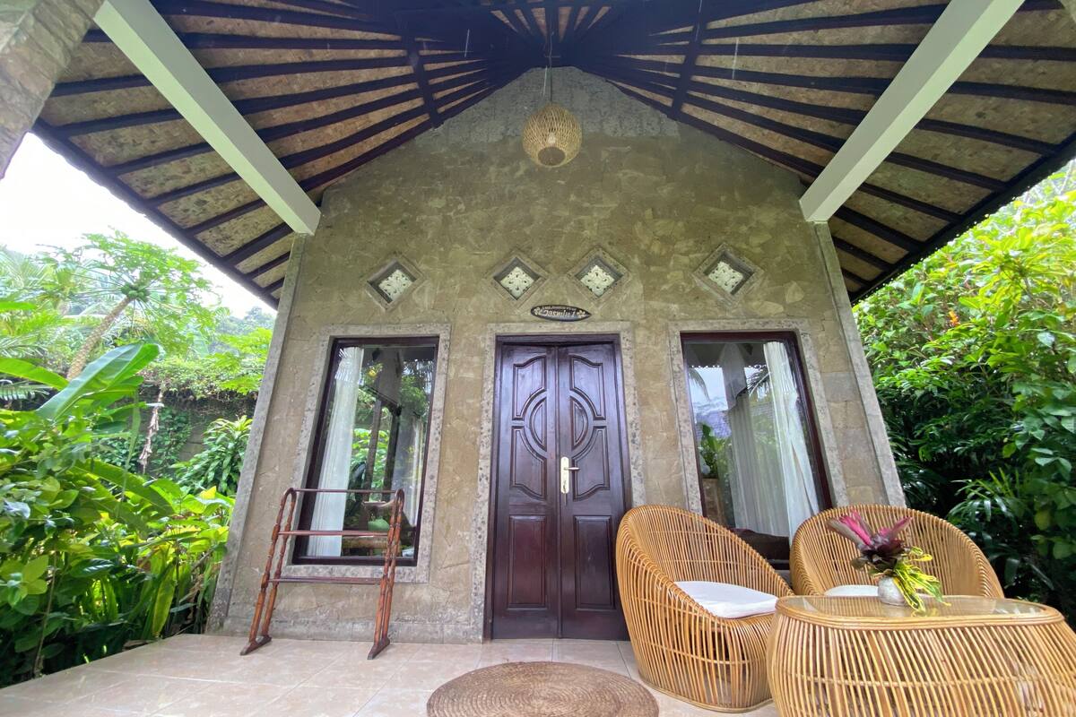 A covered porch with a wooden ceiling, a large wooden door, and two wicker chairs with a small table.
