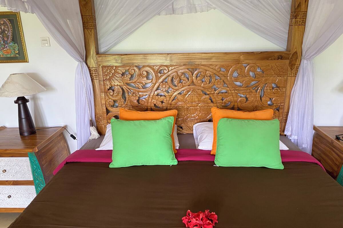 A bed with a wooden headboard, green and orange pillows, and a red flower on the bedspread.