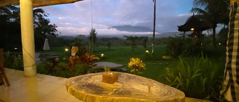 Une table en bois sur laquelle est posé un vase de fleurs, une vue sur un jardin avec une construction à toit de chaume et un ciel nuageux.