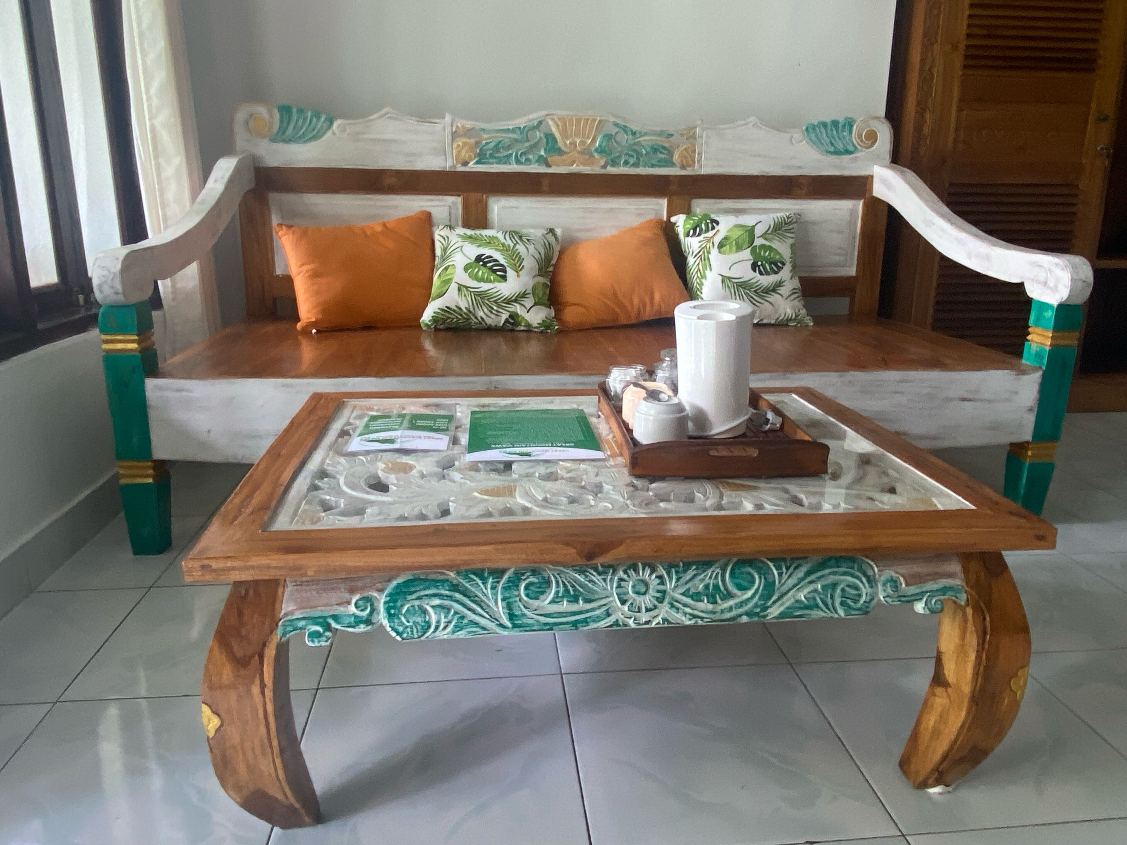 Un sofá de madera con cojines verde y naranja, una mesa de centro de vidrio con una base decorativa y una silla de madera con detalles verde y dorado.