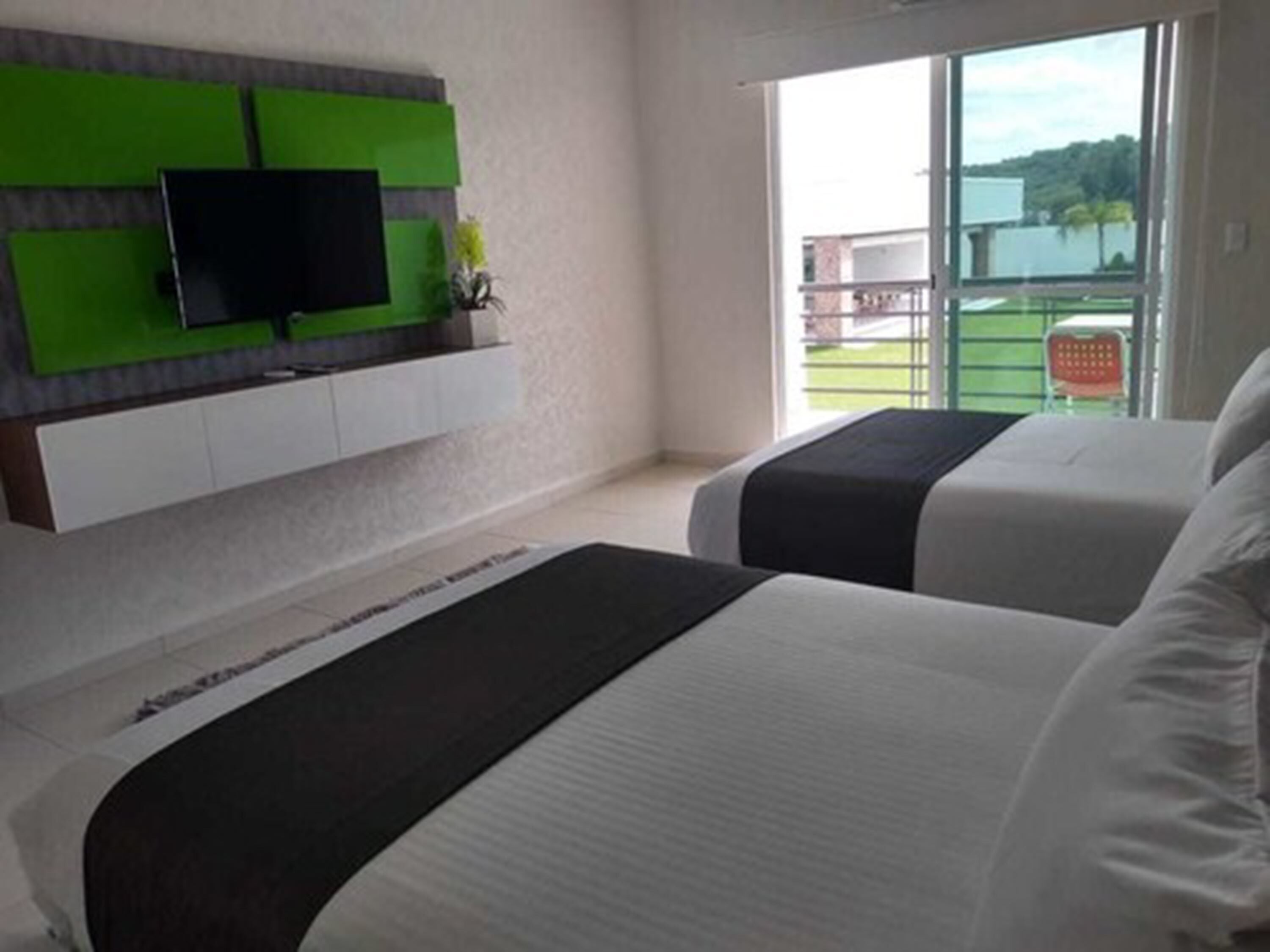 A hotel room with two beds, a flat-screen TV mounted on a green panel, and a view of a building and greenery through the window.