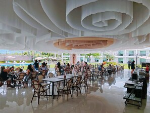 Un amplio espacio exterior con mesas y sillas, una piscina al fondo y un moderno diseño arquitectónico en el techo.