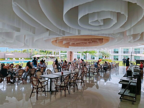Un amplio espacio exterior con mesas y sillas, una piscina al fondo y un moderno diseño arquitectónico en el techo.