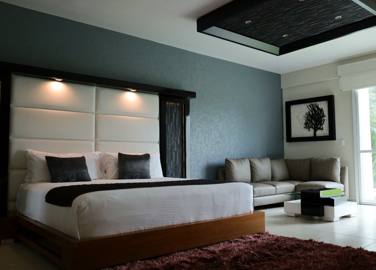 A modern bedroom with a large bed, a sofa, and a decorative tree wall art piece.