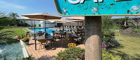 Zona de cafetería junto a la piscina con mesas al aire libre y un cartel con el nombre del establecimiento.