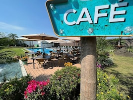 Zona de cafetería junto a la piscina con mesas al aire libre y un cartel con el nombre del establecimiento.