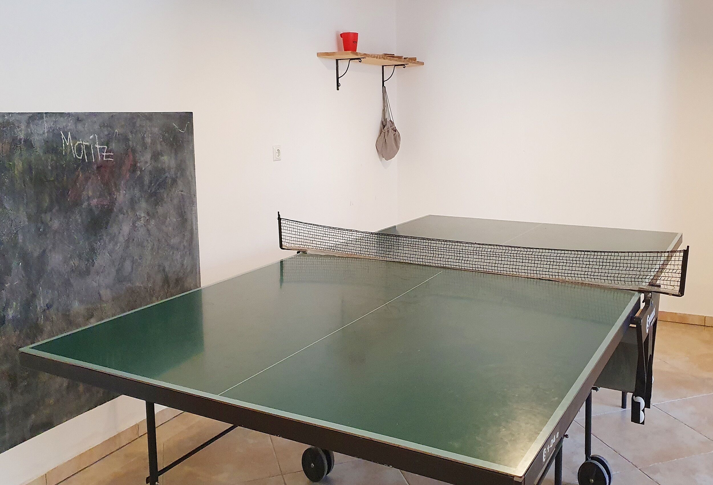 Taula de ping-pong amb rodes, pissarra amb la paraula 'Motifz' escrita, un got vermell i una bossa penjant.