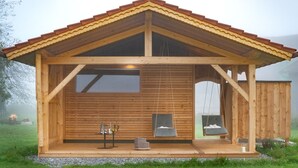 A wooden cabin with a pitched roof, large windows, a porch with seating, and a small table with items on it. - Vacation apartment Amelie, 70 sqm, balcony, 2 bedrooms (Schechen)