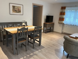 A dining area with a wooden table and chairs, a television on a wooden stand, and a window with curtains.