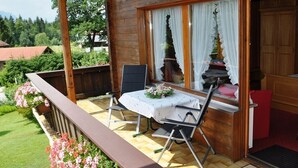 Un balcón de madera con una mesa y sillas, una puerta roja, y vistas a un jardín y edificios al fondo.