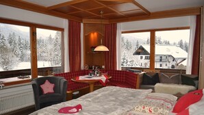 Una habitación acogedora con una cama grande, un sofá rojo a cuadros, una mesita con una lámpara y vistas a montañas nevadas a través de amplios ventanales.
