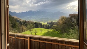 Uitzicht vanaf een balkon met houten spijlen, op een groene vallei met bomen en verre bergen onder een bewolkte lucht.
