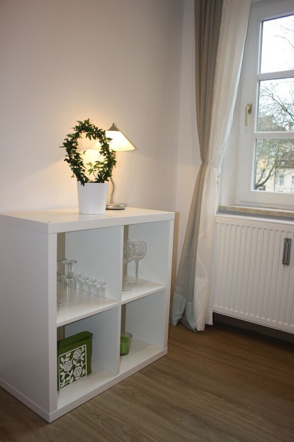 A white shelf unit with a potted plant and a lamp, a window with curtains, and a radiator.