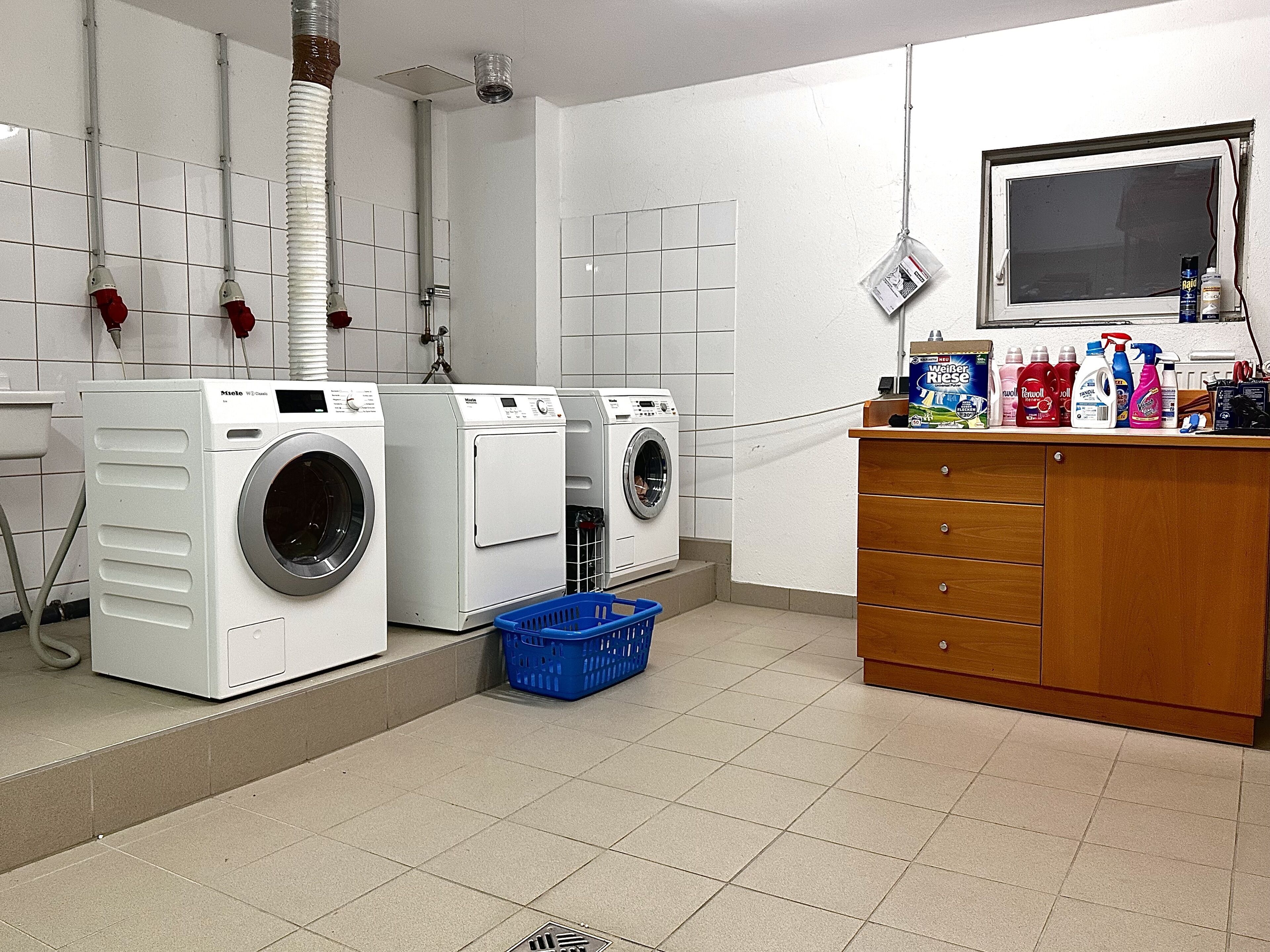 Een wasserette met twee voorladers, een blauwe wasmand, een houten kastje met schoonmaakspullen en een betegelde vloer.
