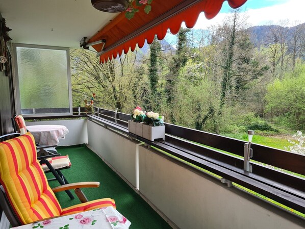 A balcony with a green artificial turf floor, a striped chair, a small table with a floral tablecloth, and a view of a forested area with mountains in the background.