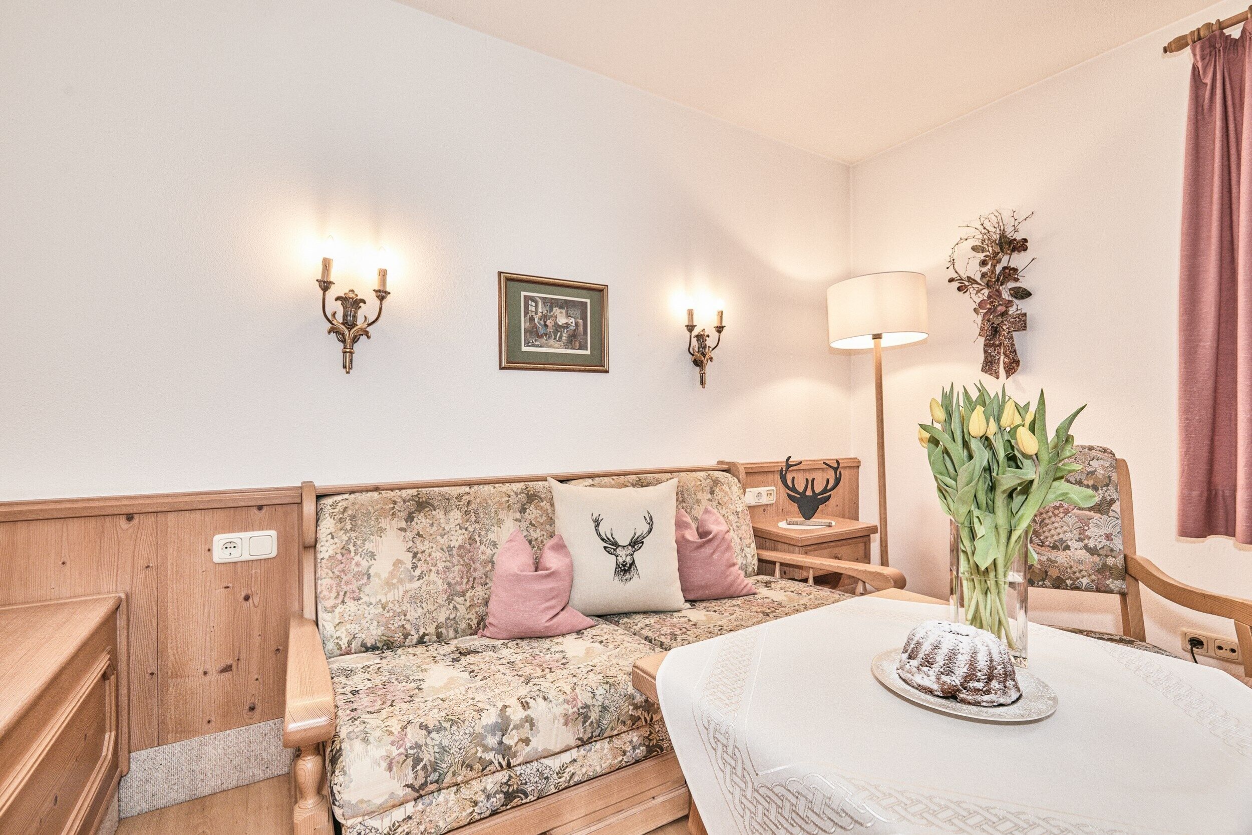 A cozy room with a floral-patterned sofa, a table with a cake, and wall-mounted lamps.