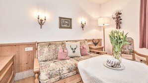 A cozy room with a floral-patterned sofa, a table with a cake, and wall-mounted lamps. - (8) Zwei-Raum-Ferienwohnung 38qm, Bad/WC, Extra-Schlafraum, Küche, Balkon (Reit im Winkl)