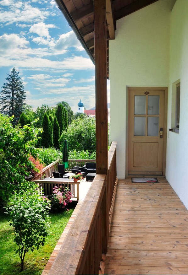Um deck de madeira com uma porta que dá para uma área de jardim, rodeado por vegetação e um céu limpo.
