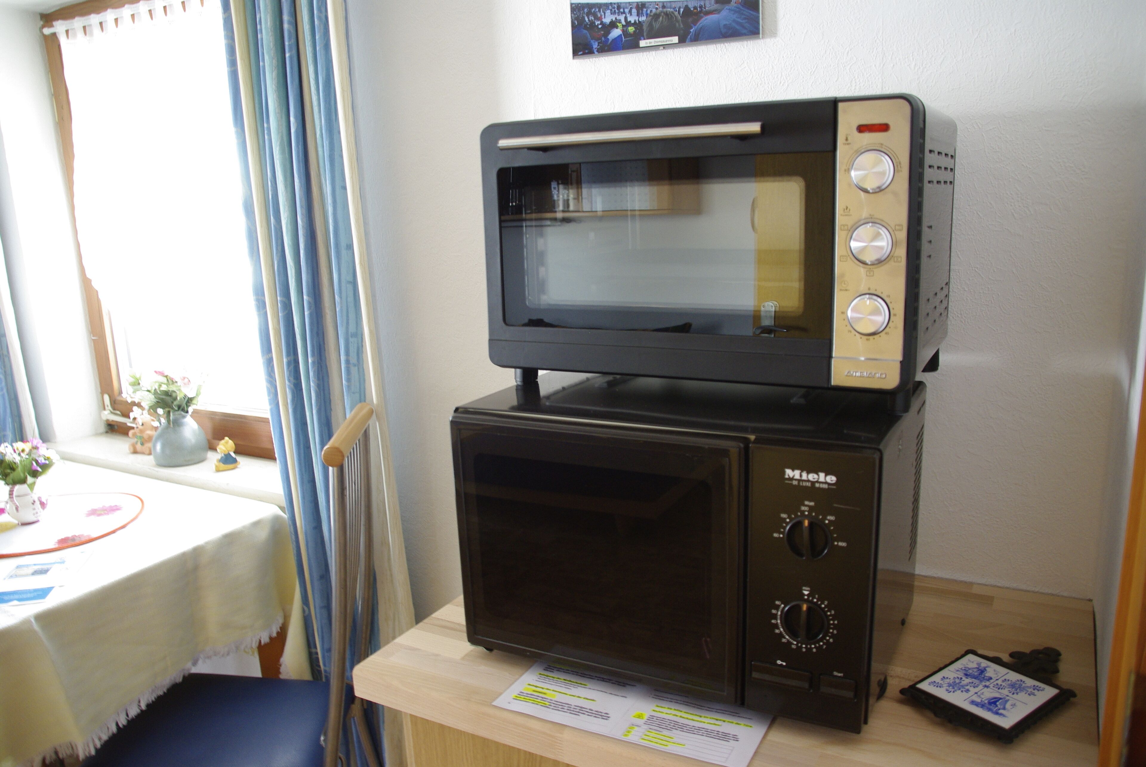 A kitchenette with a microwave oven on top of a smaller microwave, a wooden table with a white tablecloth, and a window with blue curtains.