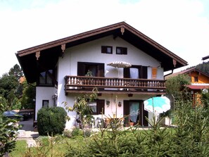 A two-story building with a balcony, a car parked in front, and a garden with trees and shrubs. - Apartment 38sqm, balcony, kitchenette, 1 bedroom, WLAN, no pets (Ruhpolding)