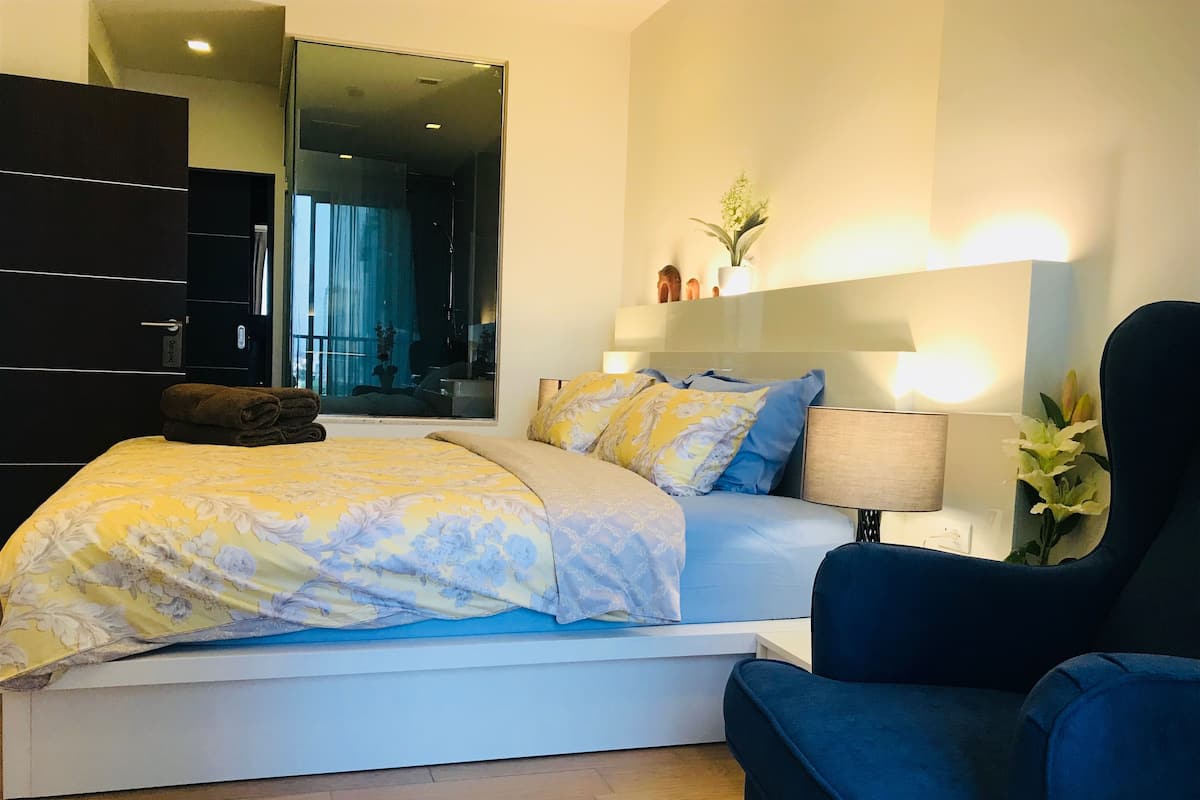 Un dormitorio moderno con una cama, un sillón azul, una mesita de noche con una lámpara y una estantería de pared con objetos decorativos.