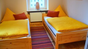 Um quarto pequeno com duas camas de madeira, cada uma com um cobertor amarelo e almofadas laranjas. Um tapete colorido listrado está no chão, e há um radiador entre as camas.