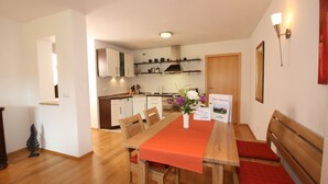 Comedor con una mesa de madera, sillas y mantel corrido rojo. Hay un jarrón con flores sobre la mesa, y un letrero con texto al fondo.