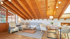 Un loft spacieux avec un haut plafond en bois, un coin salon confortable avec un canapé et des fauteuils, une table à manger avec des chaises, et de grandes fenêtres laissant entrer la lumière naturelle.