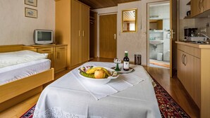 Habitación de hotel compacta con cama, un pequeño televisor, una mesa de comedor con fruta y vino, y una kitchenette.