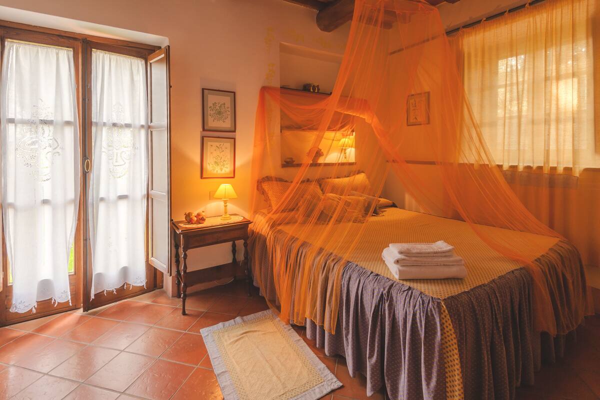 A bedroom with a bed covered in a canopy, a nightstand with a lamp, and a window with curtains.