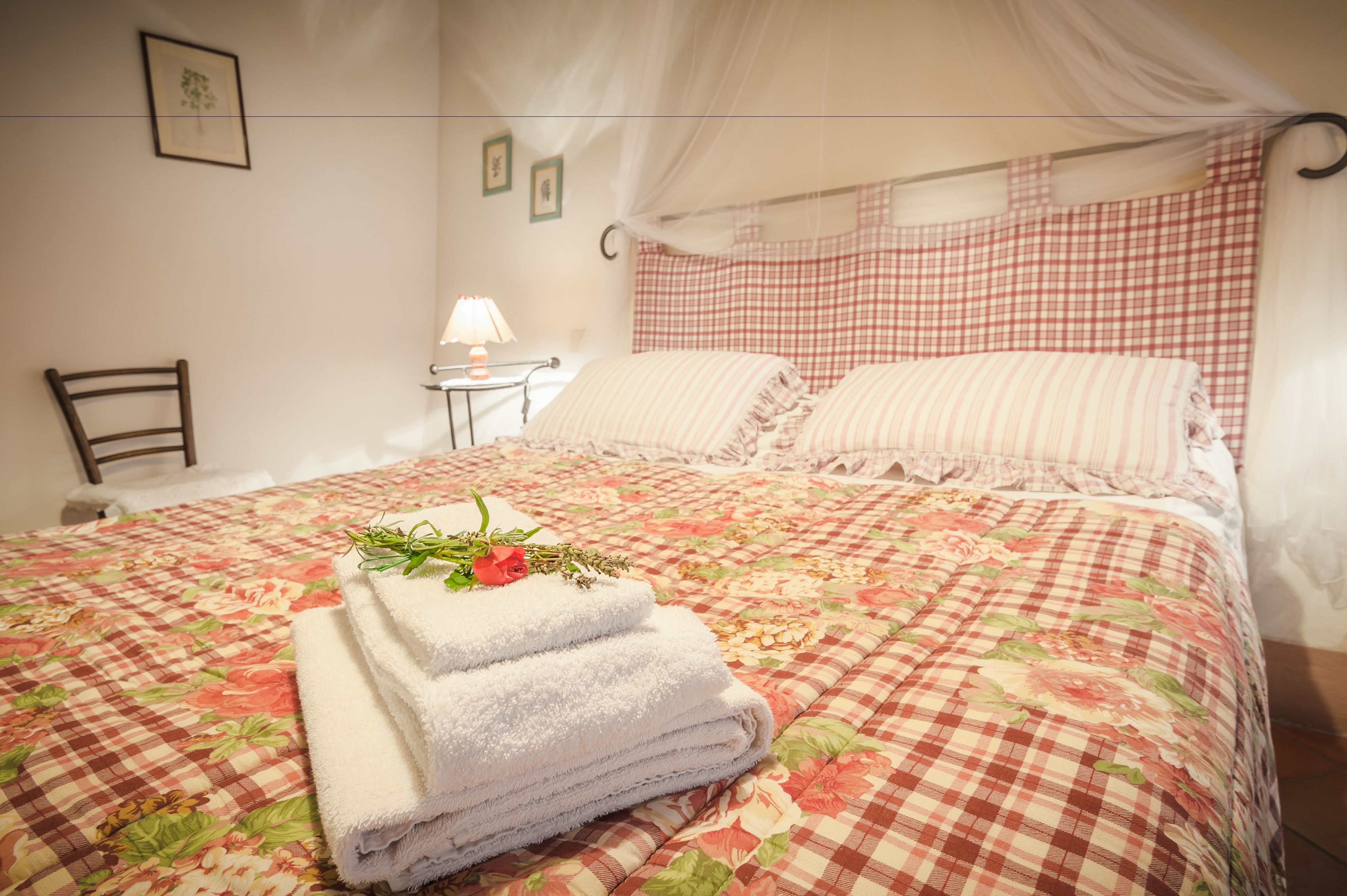 A neatly made bed with a floral bedspread, a stack of white towels, and a decorative arrangement of greenery and a rose.