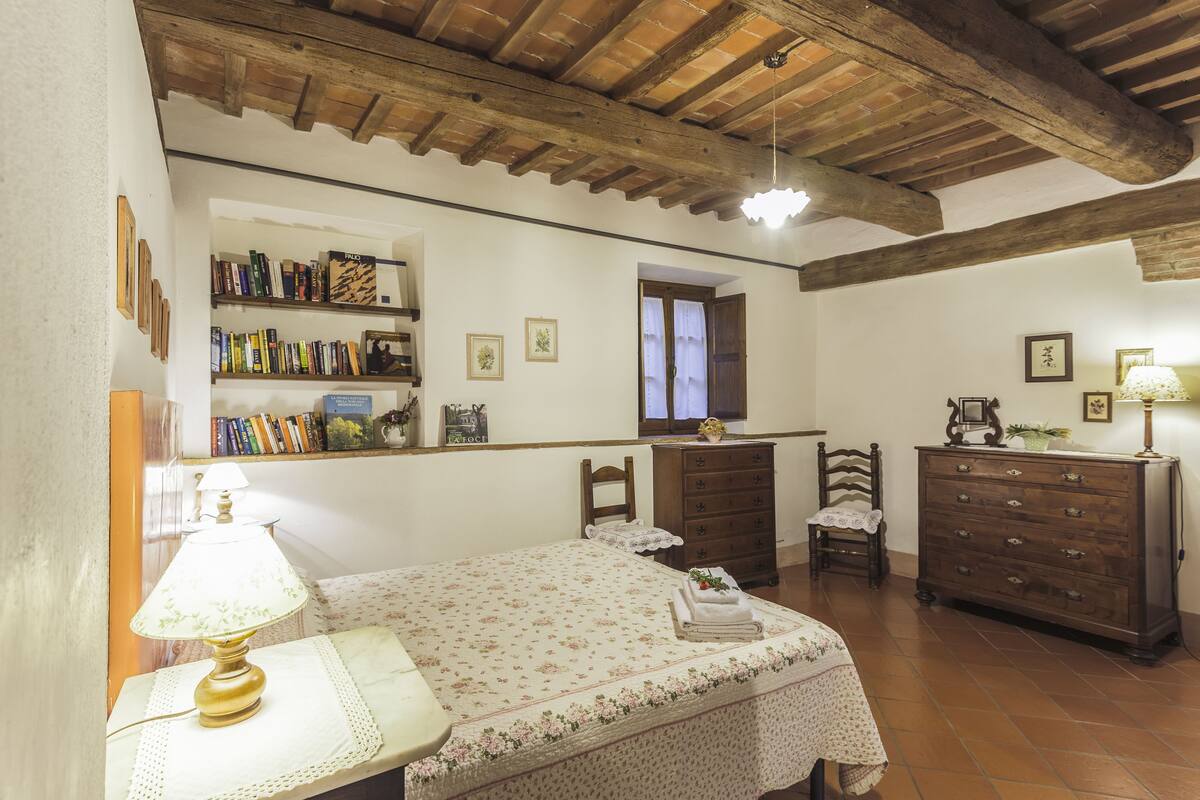 A bedroom with a wooden ceiling, a bed with a floral bedspread, a dresser, a chair, and a bookshelf with books and framed pictures.