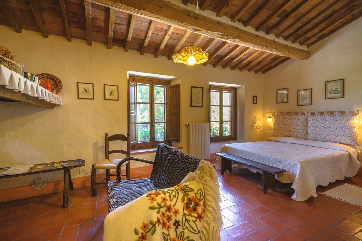 A cozy room with a wooden ceiling, a bed with a white bedspread, a chair with a floral pillow, and a table with a menu.