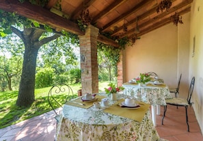 A covered outdoor dining area with tables set for two, surrounded by greenery and a large tree.