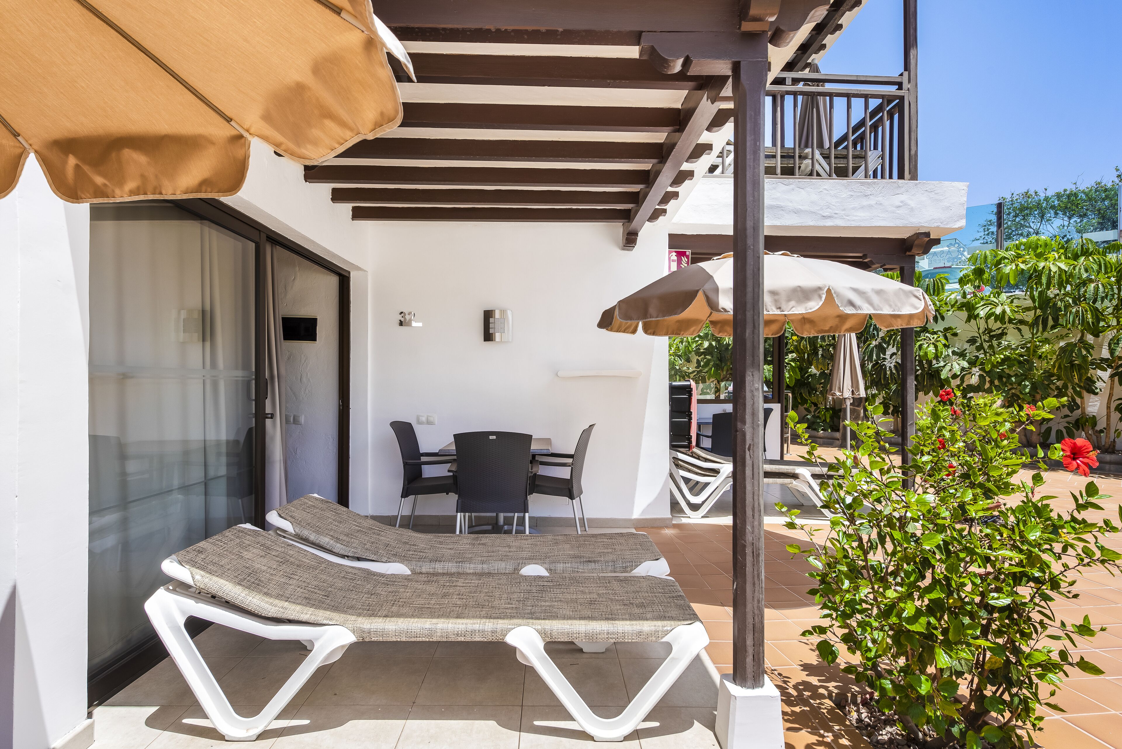 Eine Terrasse mit Liegestühlen und Sonnenschirmen, ein Essbereich mit Tisch und Stühlen sowie ein kleiner Garten mit Pflanzen und Blumen.