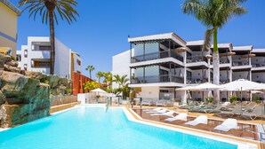 A swimming pool with lounge chairs and umbrellas, surrounded by modern buildings with balconies and palm trees. - Casas Pepe Apartments & Spa- Adults Only (San Bartolomé de Tirajana)