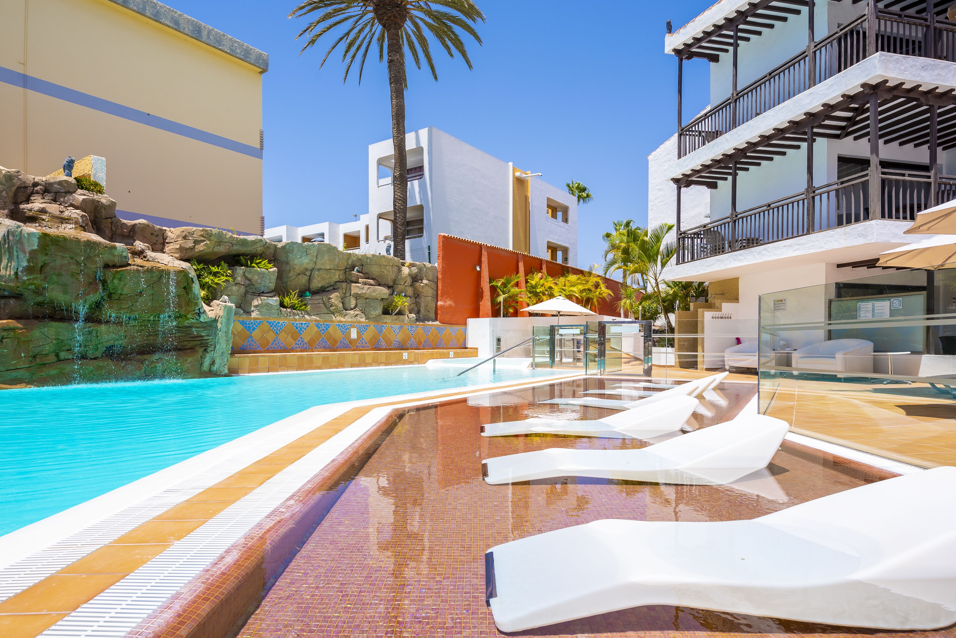 Una piscina con sillones de descanso, una palmera y edificios modernos al fondo.
