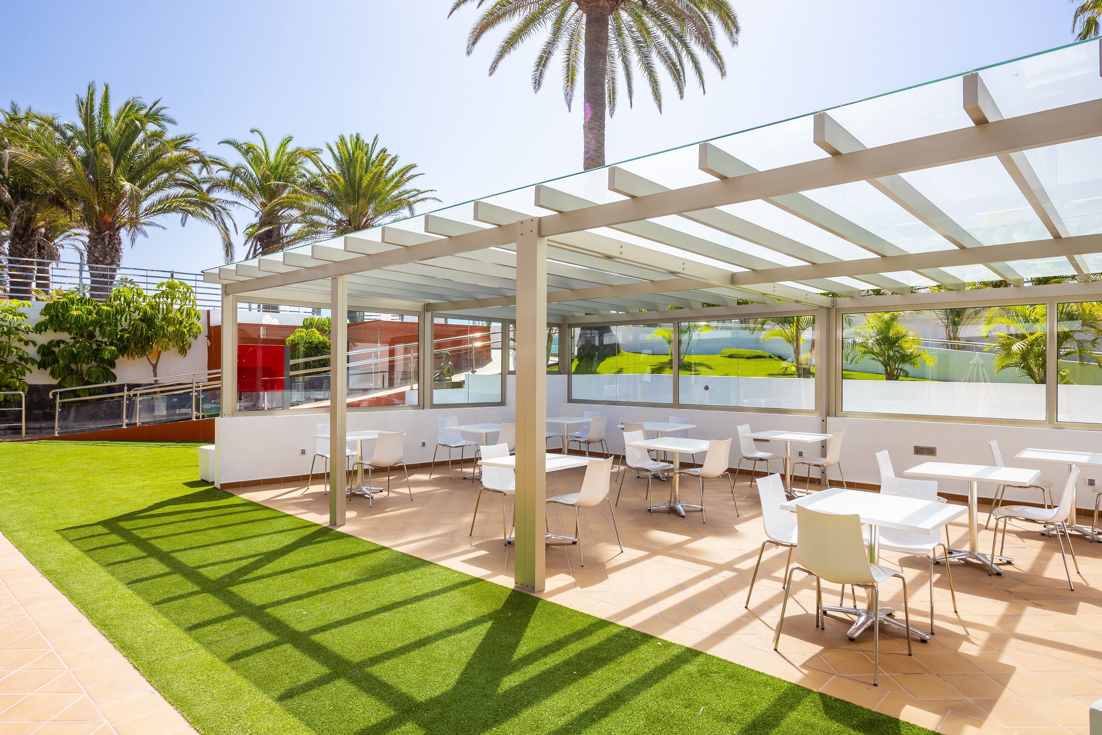 Patio esterno con tavoli e sedie bianche, circondato da pannelli di vetro e palme.