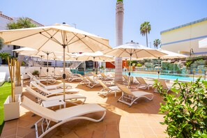 Zona junto a la piscina con tumbonas y sombrillas, piscina y una chimenea alta al fondo.
