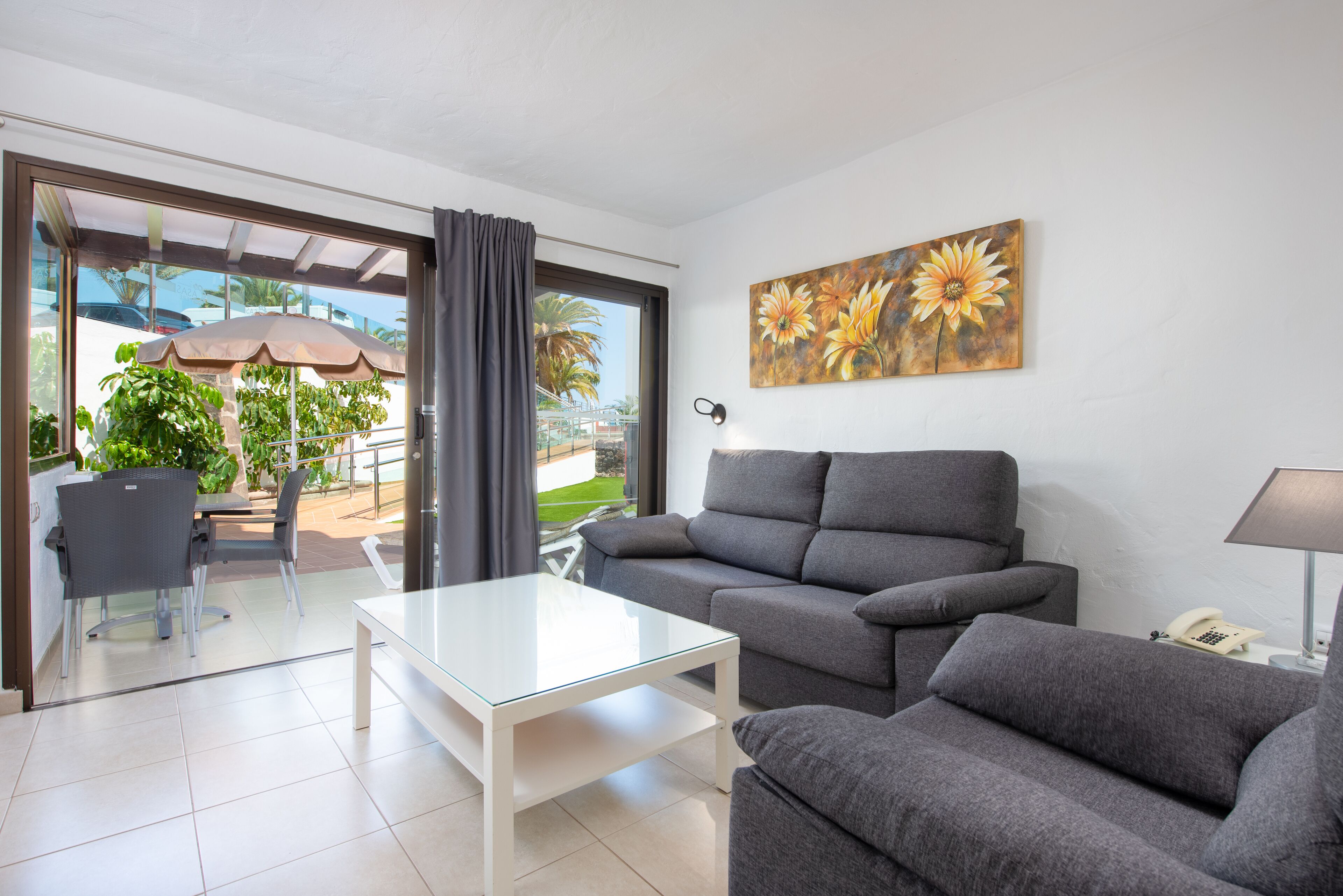 A modern living room with a grey sofa, a glass coffee table, and a dining area with chairs and an umbrella.
