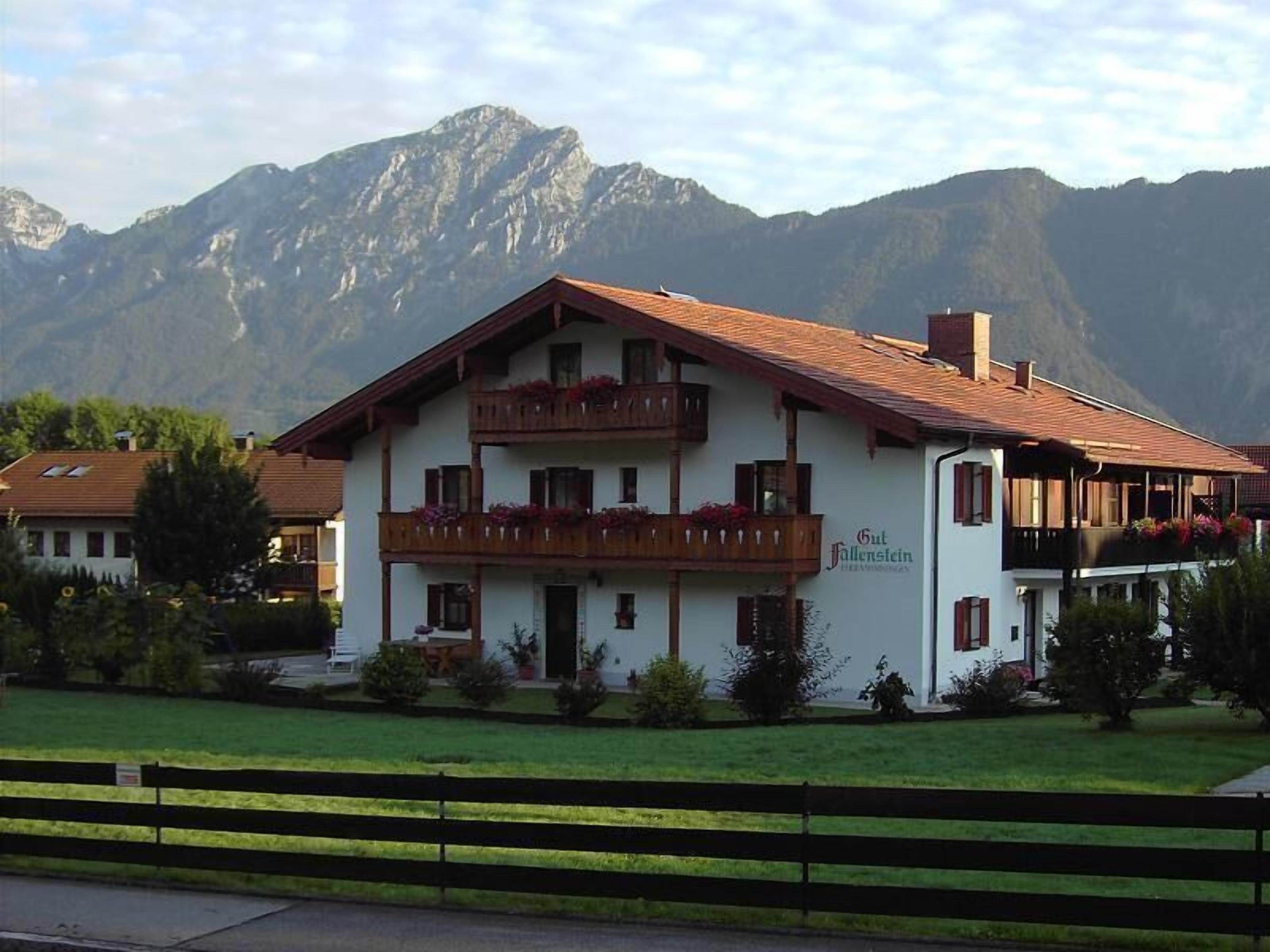 Un edificio de dos pisos con un balcón, rodeado de césped y montañas al fondo.