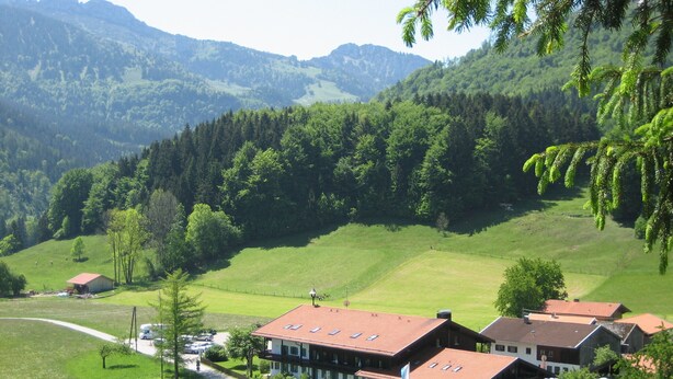 Un grand bâtiment doté de plusieurs toits, entouré de champs verdoyants et d’arbres, avec des montagnes en arrière-plan.